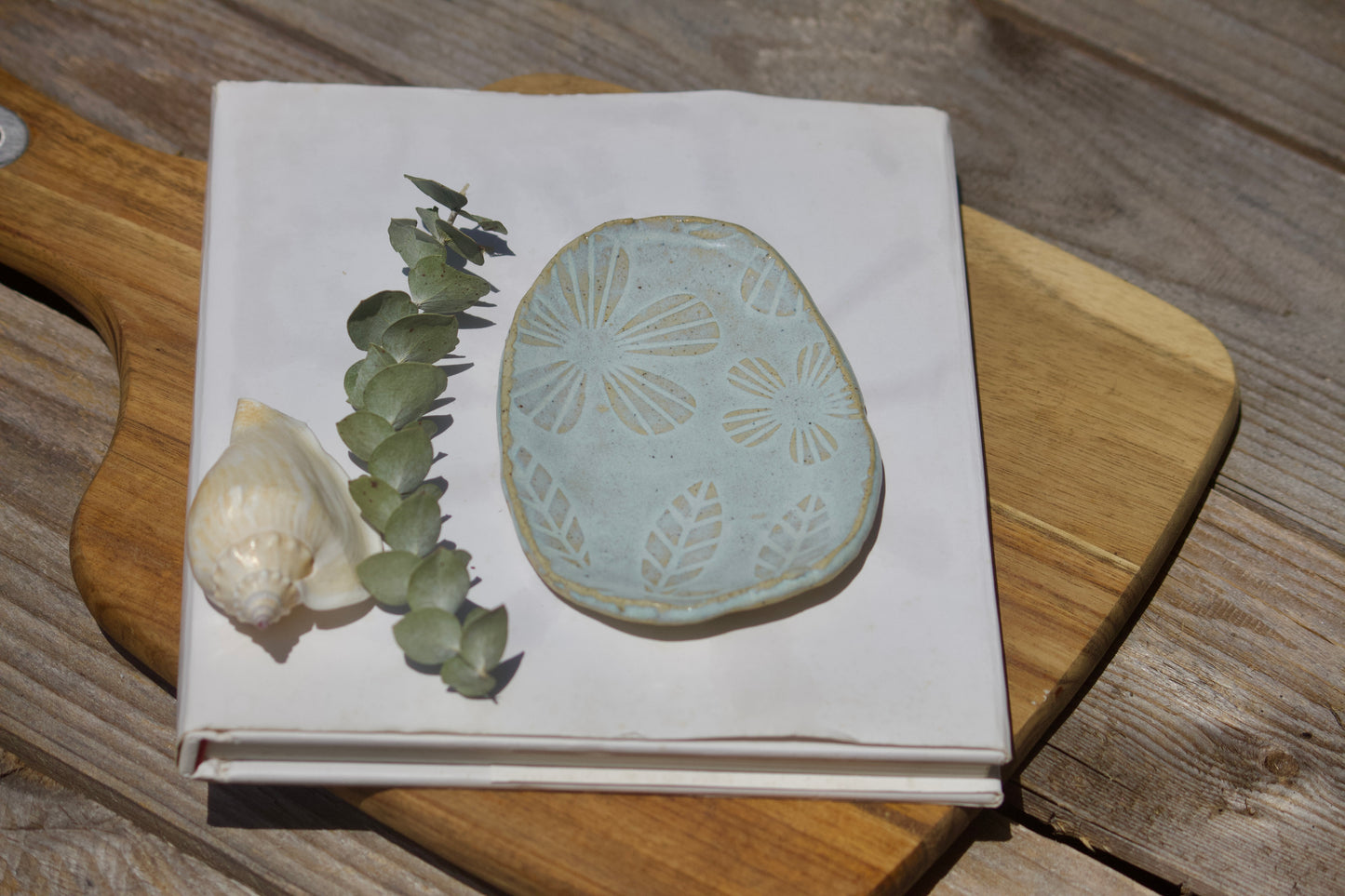 Blue oval ceramic trinket plate with floral design dislayed on white notebook on top of a wooden board with a shell next to it.
