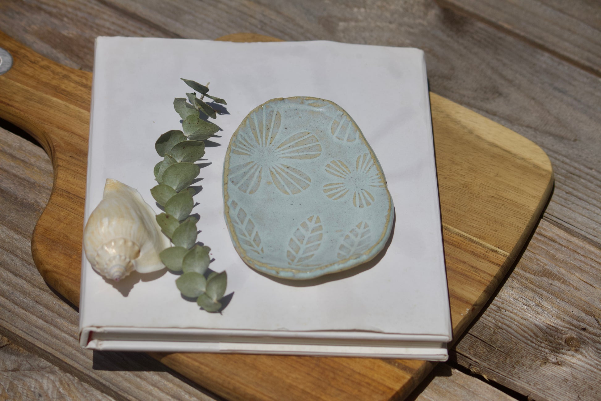 Blue oval ceramic trinket plate with floral  design dislayed on white notebook on top of a wooden board with a shell next to it.