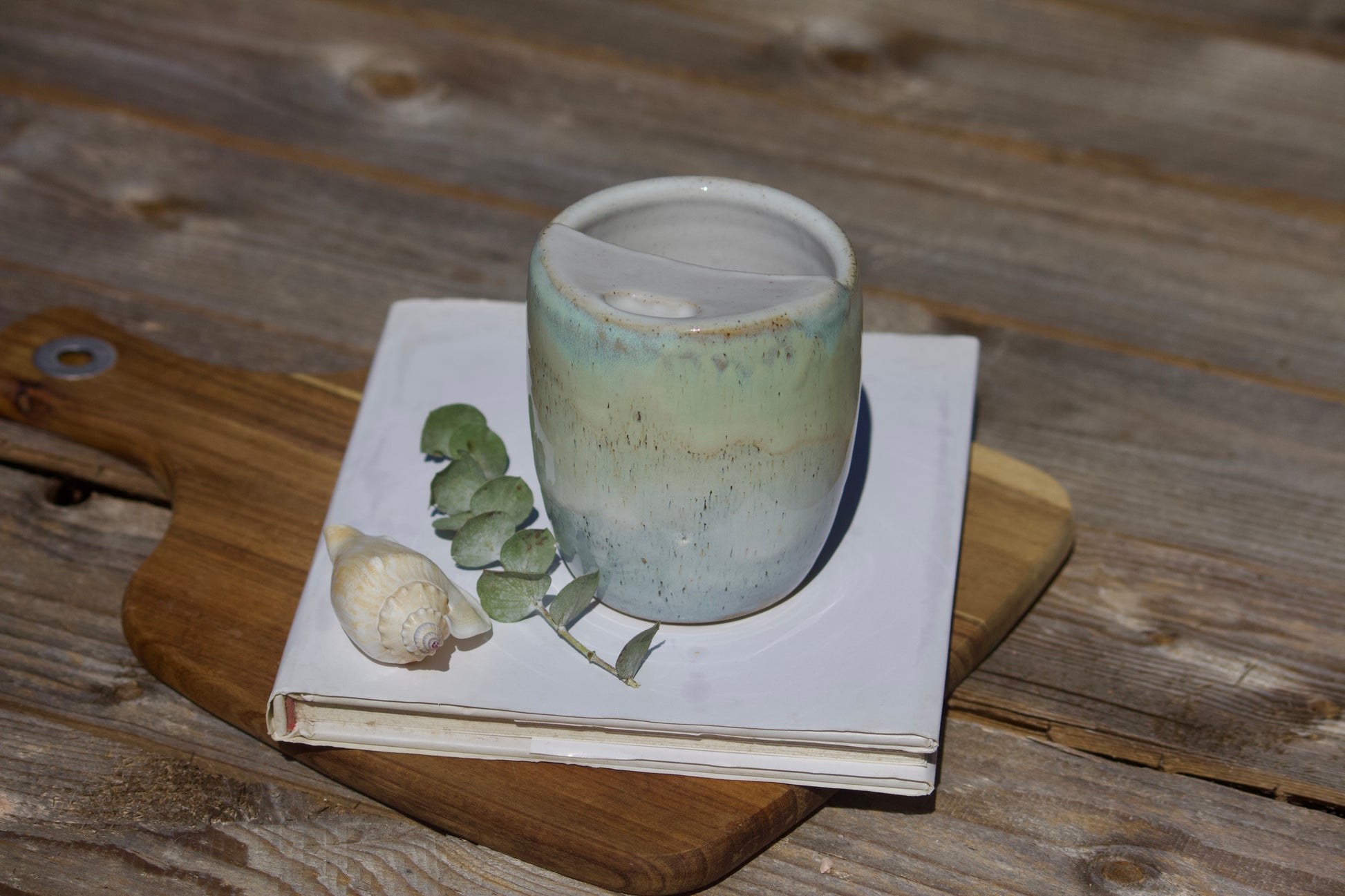 A ceramic travel mug with a mix of green and blue colors, placed on a white notebook, accompanied by a small decorative item and a leaf on a wooden surface.