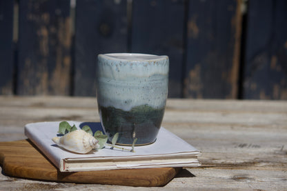 A ceramic travel mug with a mix of light and dark blue coluors, placed on a white notebook, accompanied by a small decorative item and a leaf on a wooden surface.