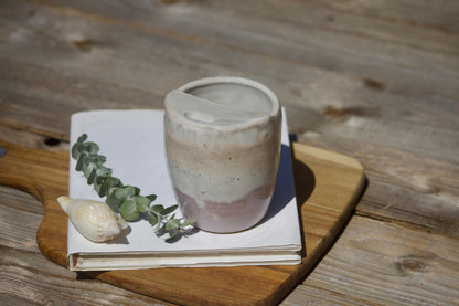 A ceramic travel mug with a mix of beige and pink colours, placed on a white notebook, accompanied by a small decorative item and a leaf on a wooden surface.