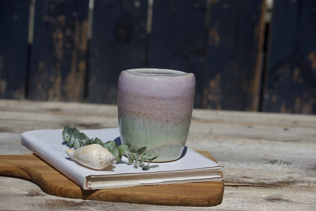 A ceramic travel mug with a mix of pink and green colours, placed on a white notebook, accompanied by a small decorative item and a leaf on a wooden surface.