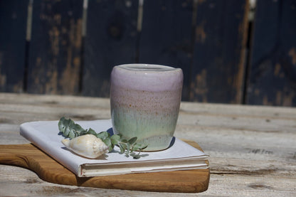A ceramic travel mug with a mix of pink and green colours, placed on a white notebook, accompanied by a small decorative item and a leaf on a wooden surface.