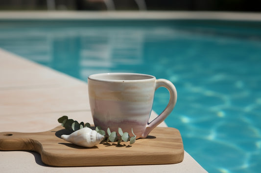 Australian handmade ceramic pastel ceramic cup with pink and peach hues, sitting on top of a stack of books with a small decorative object to the side, on a wooden surface.