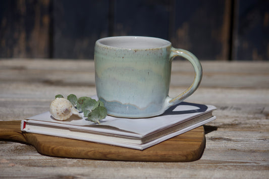 A seafoam pastel green and blue ceramic mug placed on top of a closed journal, which is on a wooden table. There is also a small decorative item and a leaf near the mug.