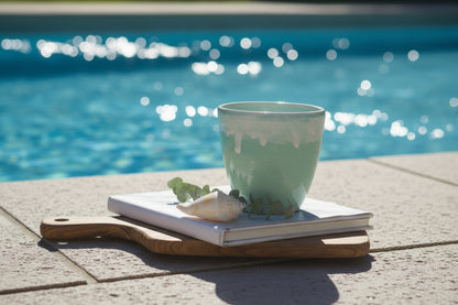 A turquoise ceramic cup with a handleless design, placed on a wooden board next to a wooden notebook and a shell, with a black background.
