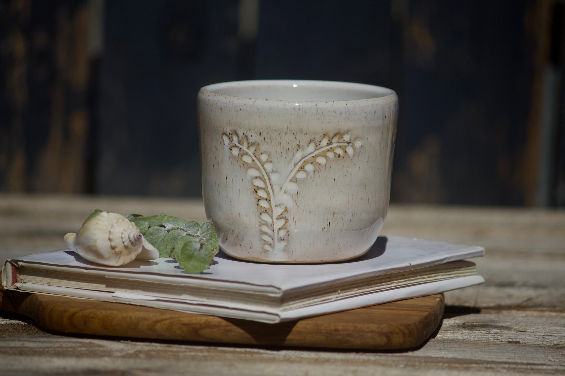 A carved wheat ceramic cup with a leaf design, placed on a wooden tray next to a shell and some green leaves, with a blurred background.