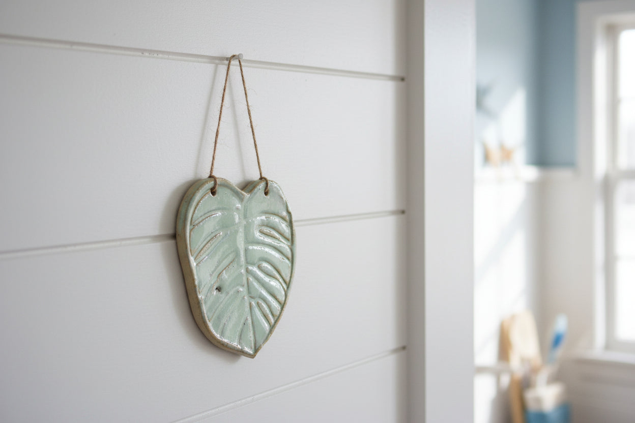 A green leaf-shaped ceramic wall hanger, made from Australian clay, displayed on a wooden board with a white fabric and some beach accoutrements.