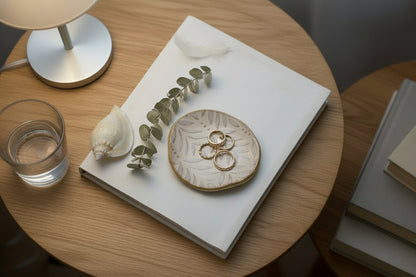 A round beige leaf ceramic trinket plate with a natural leaf carving design, displayed on a wooden surface, accompanied by a wooden box and some dried flowers.